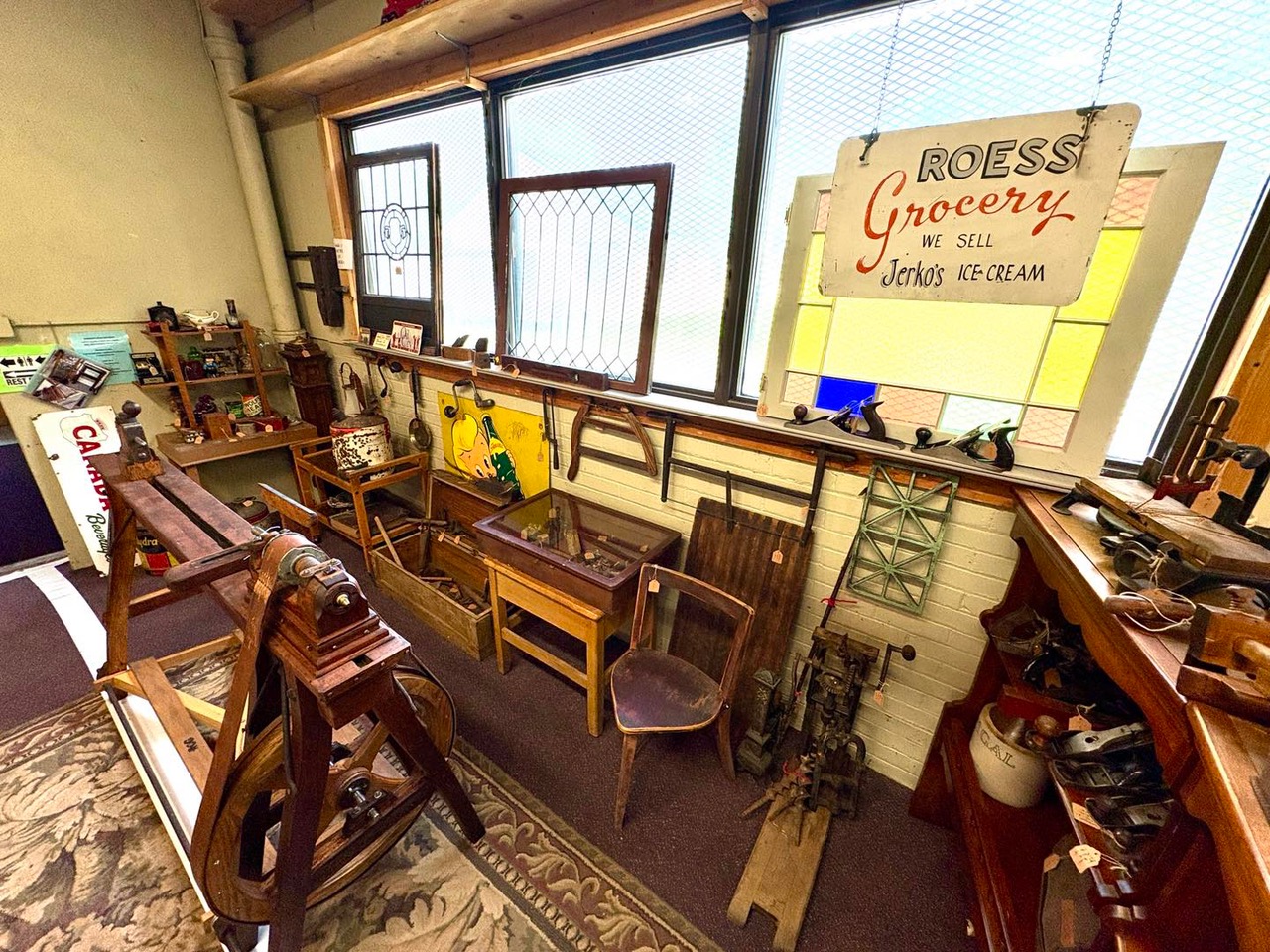 Eclectic vintage tool and antique display featuring a Roess Grocery sign, stained glass, hand tools, and rustic woodworking equipment—perfect for collectors and nostalgia lovers
