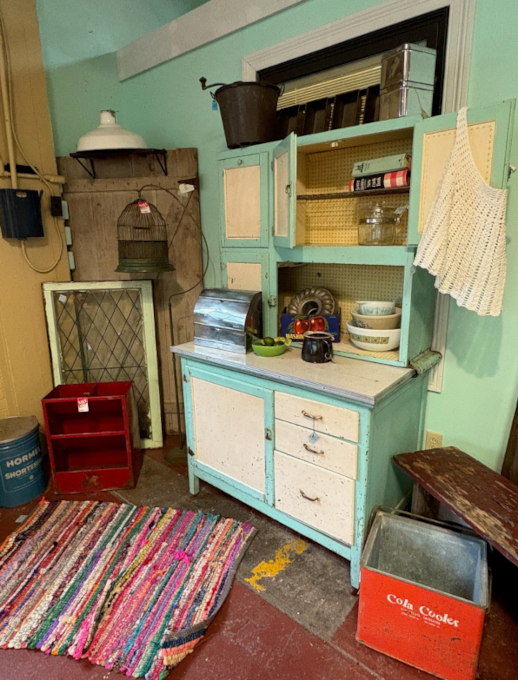 Vintage kitchen setup with mint green Hoosier cabinet, retro cookware, antique birdcage, red shelf, metal cola cooler, and multicolored rag rug in a rustic home décor scene.