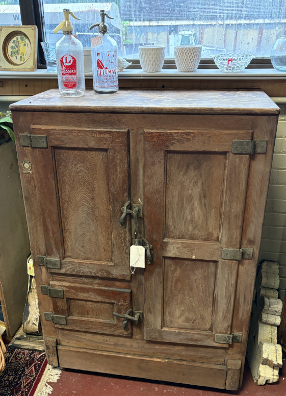 Antique wooden icebox with metal latch hardware and aged patina, featuring multiple compartments for vintage cold storage, displayed with collectible glass seltzer bottles on top