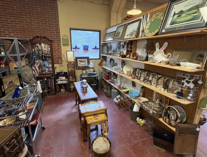 Charming antique shop corner showcasing a curated mix of vintage décor, collectible plates, framed artwork, glassware, and patriotic accents, all beautifully arranged on rustic wooden shelves and tables for a nostalgic shopping experience.