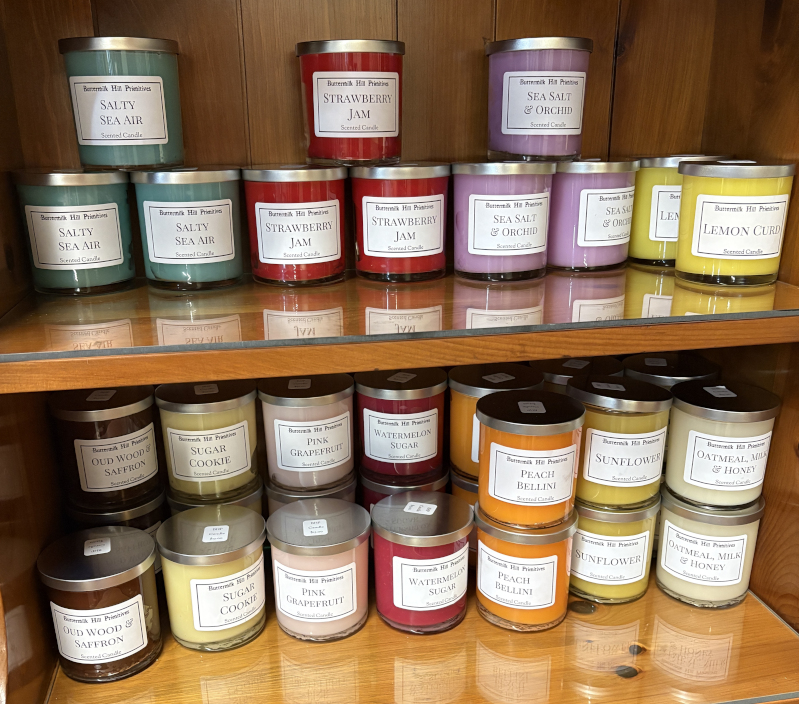 Colorful assortment of scented candles on wooden shelves, featuring fragrances like Salty Sea Air, Strawberry Jam, Sea Salt & Orchid, Lemon Chiffon, Oud Wood & Saffron, Sugar Cookie, Pink Grapefruit, Watermelon Sugar, Peach Bellini, Sunflower, and Oatmeal, Milk & Honey.