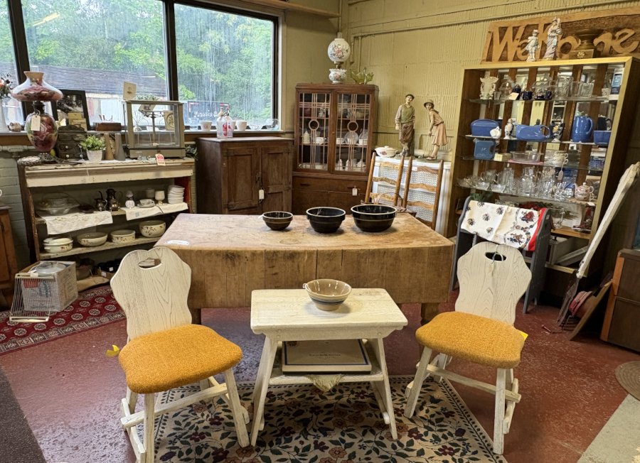 Eclectic antique shop display featuring vintage furniture, mid-century chairs with orange cushions, rustic wooden butcher block table, collectible bowls, and shelves filled with retro dishware and glassware.