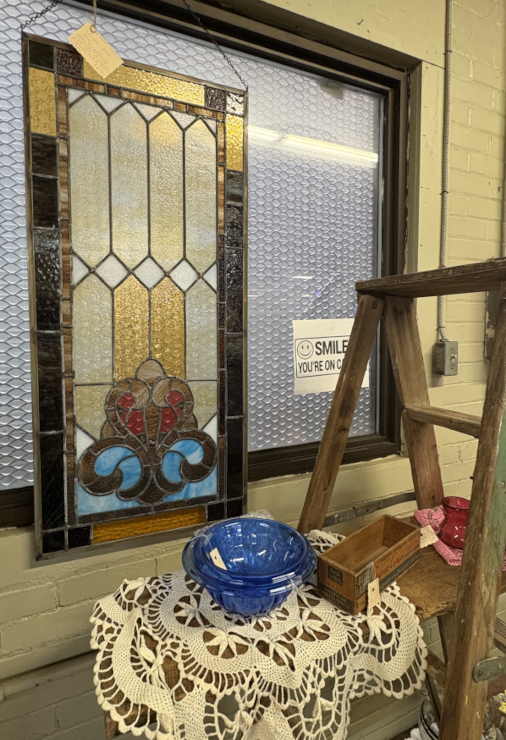 Vintage stained glass window panel with colorful geometric design, displayed alongside a crochet tablecloth, blue glass bowl, and rustic wooden ladder in an antique shop setting.