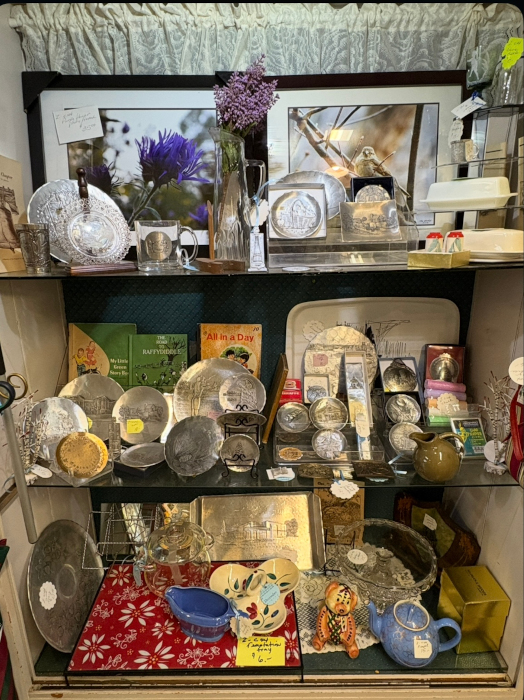A display cabinet filled with vintage pewter plates, bowls, and trays, along with books, framed photos, glassware, and small decorative items.