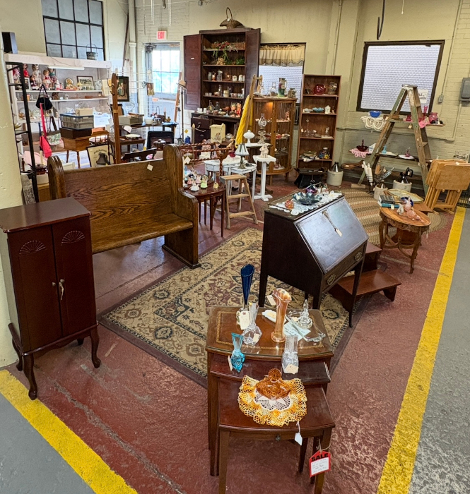 An antique shop section with wooden furniture, glass vases, small tables, and shelves filled with vintage decor, arranged on patterned rugs.